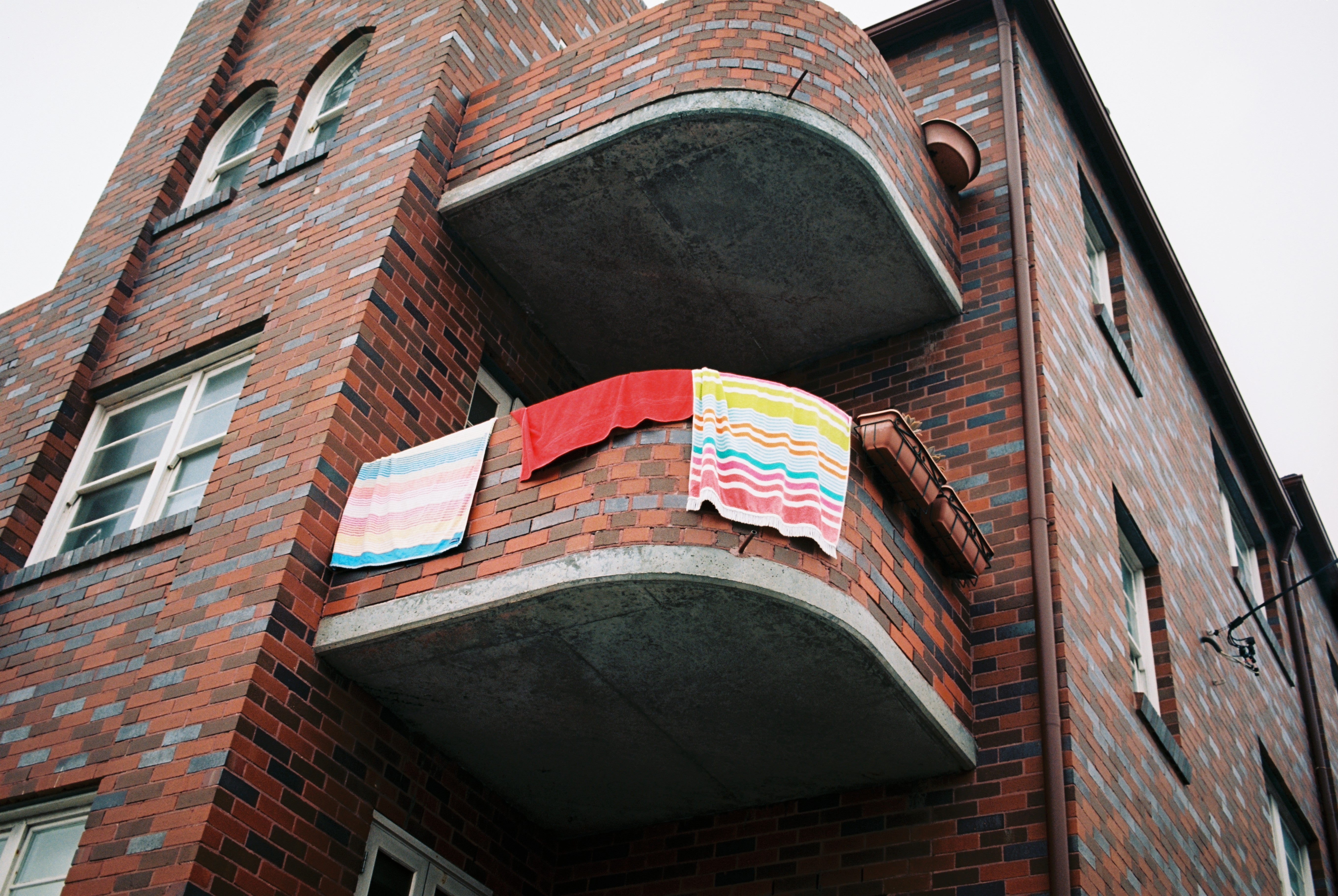 A photography of coloured beach towels hanging over the balcony of an old red brick apartment building in Bondi, Sydney Australia