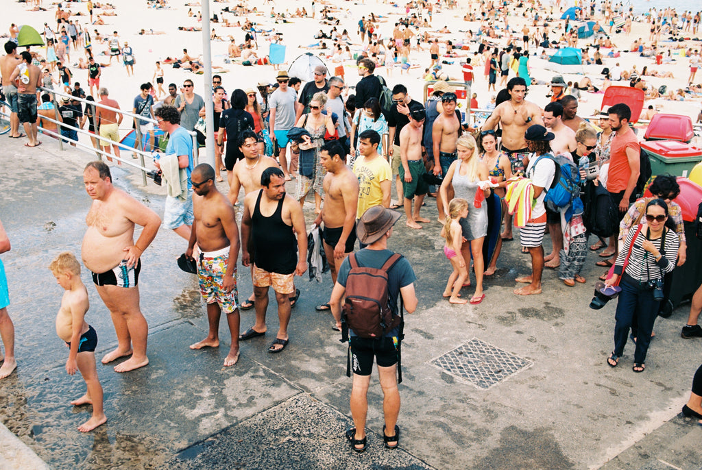 “A photo of New Years day at Bondi Beach, Thousands of people on the beach & in the water. People cue to have a shower in a long line. People are wearing bathers, it’s a hot day in Sydney Australia.