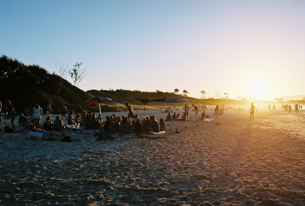 People sit in groups on the beach at sunset in Byron bay. The sun is low & the beach & blue water looks warm.  People are walking in the distance.