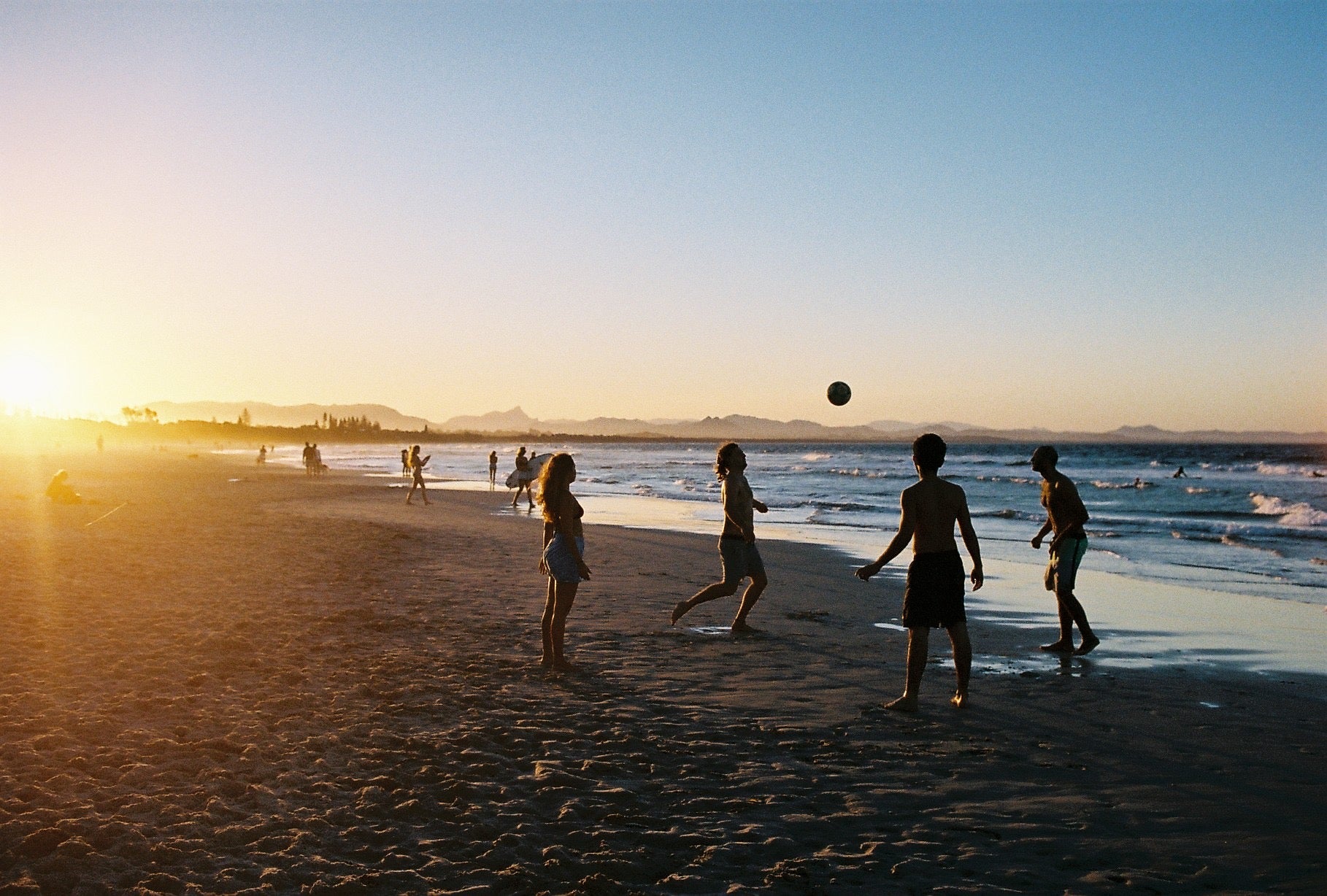 A room with a photograpic framed picture on the wall. There is a wooden seat under the picture. Young people play soccer on the beach at sunset in Byron bay. The sun is low & the beach & blue water looks warm. 