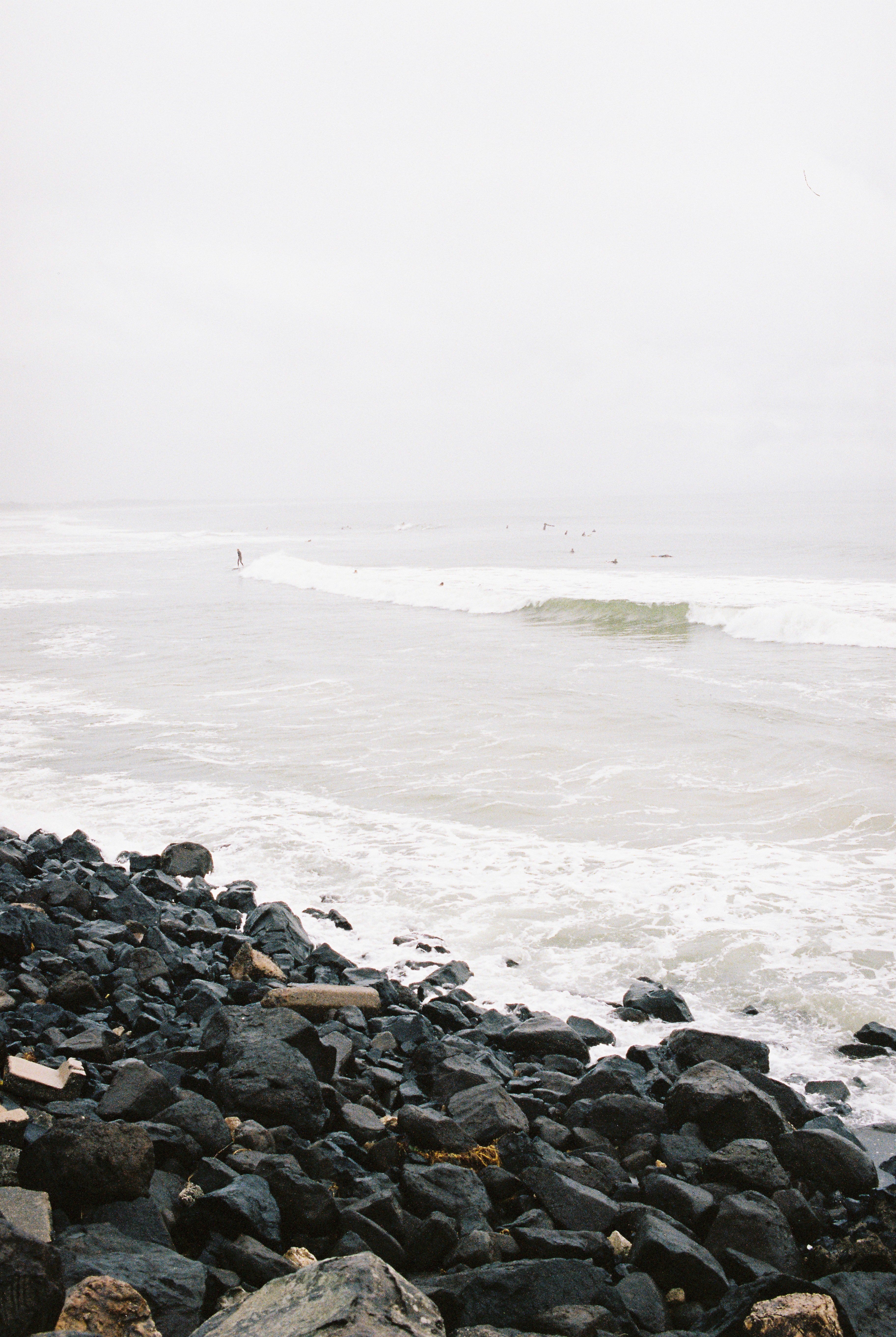 A photograph of waves breaking in Byron bay.  the water is white & hazy, white clouds. There are surfers in the water & rocks in the foreground. Main Beach, byron bay, 