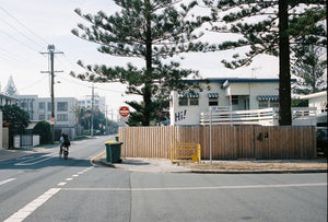 A colour photograph of an old beach house on the corner of a quiet street near the beach on the Goldcoast of Australia. There is a young man on his push bike on the street riding. Artwork Prints, wall art, Melbourne travel, Australia Photographic prints, Framed artwork, Australian Photography, Film photography, Vintage photo, style Interior design, Pictures, Abstract, film photography, photography for sale, music festival, architecture, gold coast, architecture, urban photography,