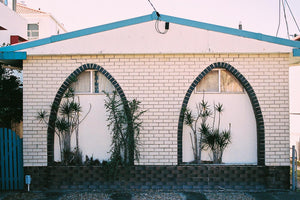 A photograph is a 1960’s brick retro looking home. Its has two arches with black bricks in a pattern around the arches. There are small plants between the arches. Artwork,