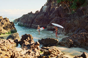 Two young girls walk into the water around the rocks to go surfing. They are wearing bathers and carry surfboards. The blue water looks warm.