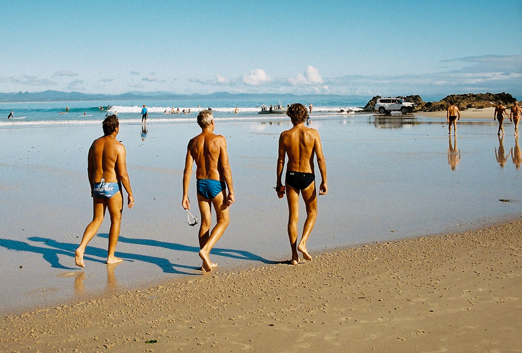 Three local men in speedos walk along Byron Bay's clarkes beach ready to go for a swim