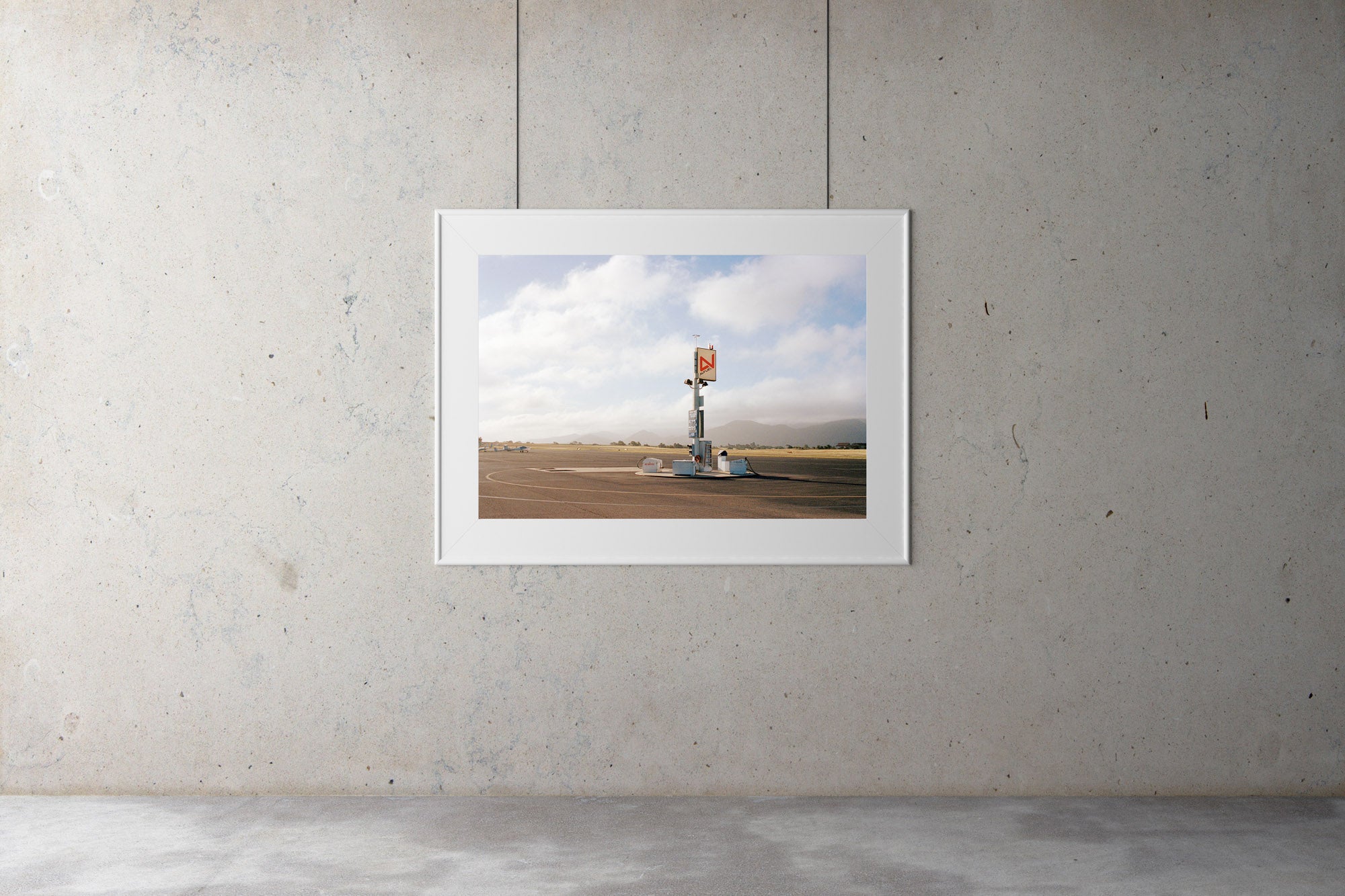 A photographic print gallery with an image on the wall. A photograph of an old airfield with a petrol filling station & old airplanes in the photo. Blue skies & clouds in the background. USA