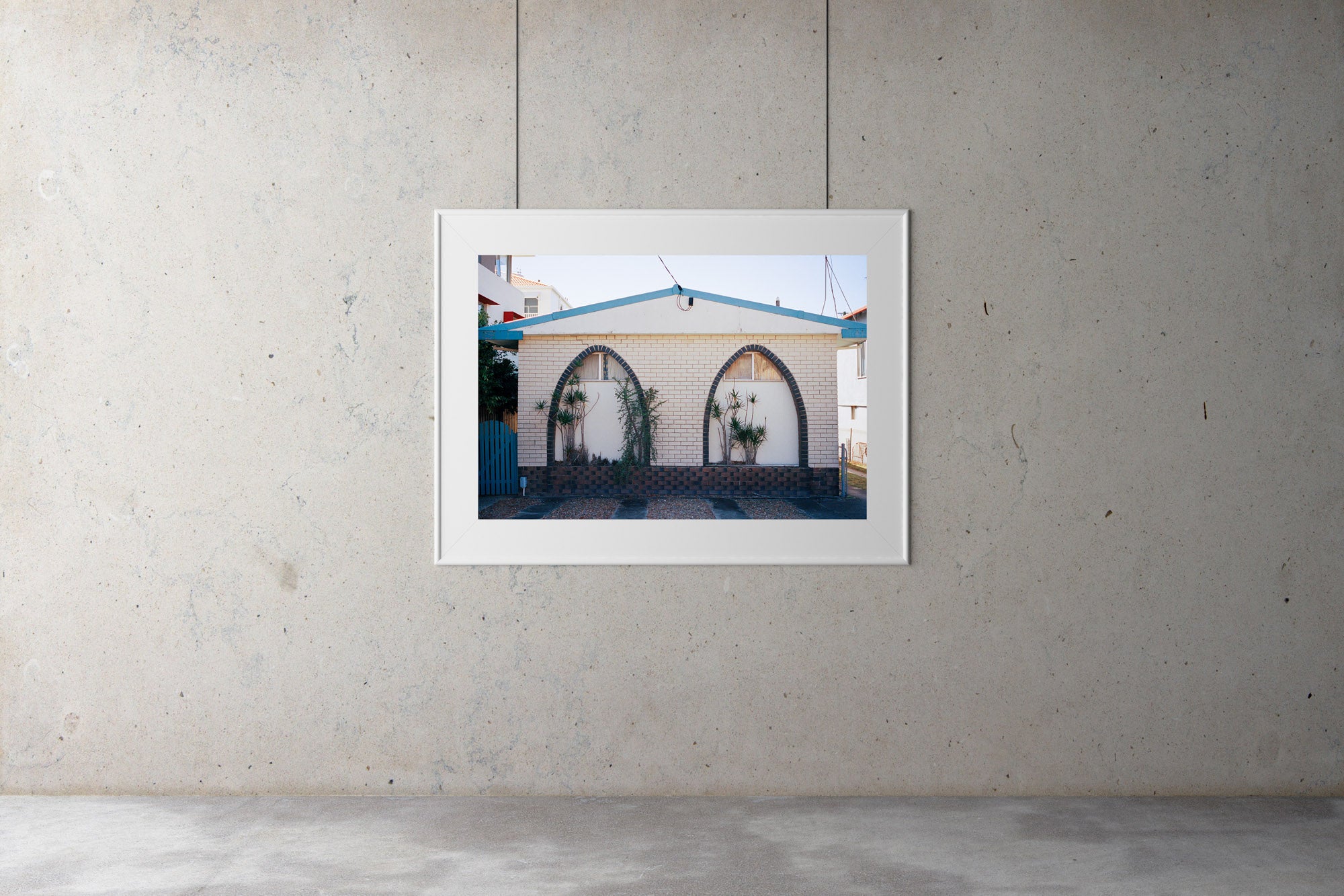 A photograph in a gallery.A photograph is a 1960’s brick retro looking home. Its has two arches with black bricks in a pattern around the arches. There are small plants between the arches. Artwork,
