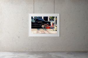 A photograph of a red bike leaning on a pole in a city street in Tokyo. There is a black taxi in the street.  Tokyo Japan. 