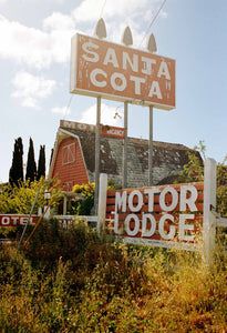 A photograph of an old 1950's American Motel. Large signs out the front & overgrown vegetation. The Motel is Barn style.