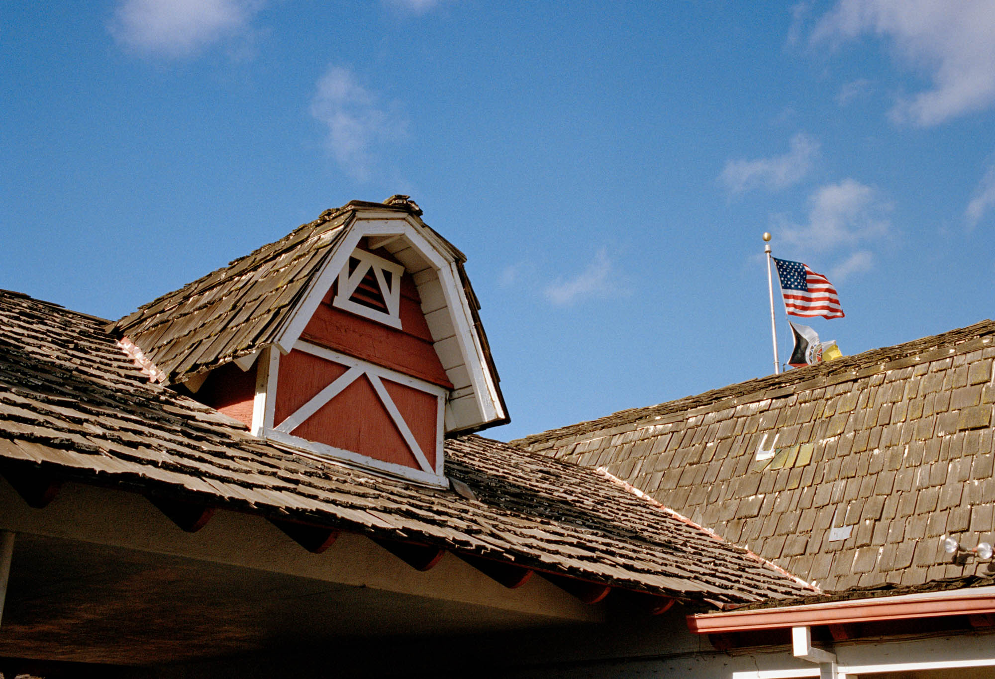 A vintage motel with shingle roof tiles & farmhouse barn roof features. There is an American flag on the roof. There are blue skies.