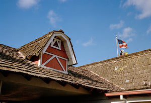 A vintage motel with shingle roof tiles & farmhouse barn roof features. There is an American flag on the roof. There are blue skies.