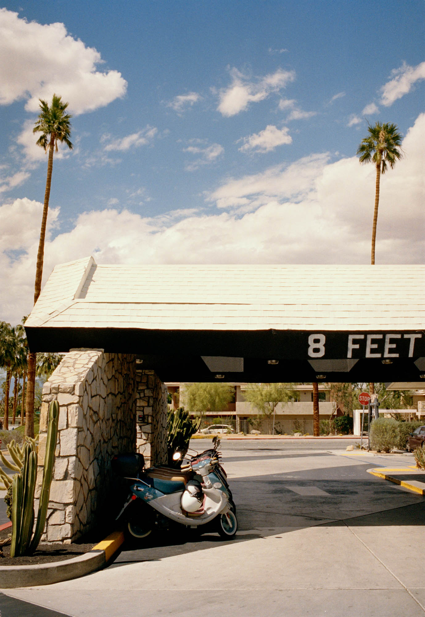 A photograph of a mid century motel in Palm Springs. There is an entrance with stone walls wth old scooters parked outside, there are palm trees & blue skies.