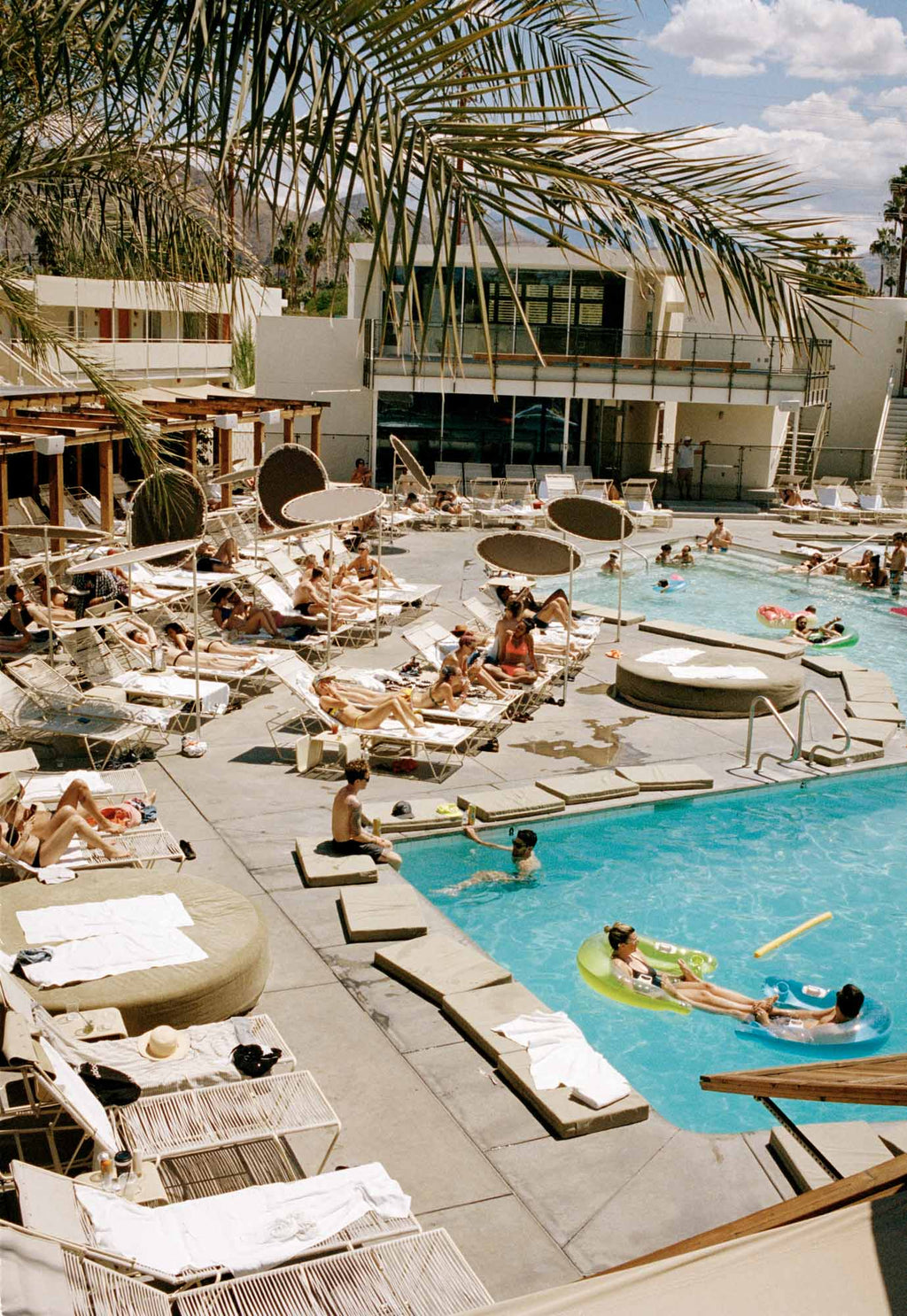 People lay around a 1950’s style Motel swimming pool on a hot day in Palm Springs California. Blue sky with clouds & palm trees.