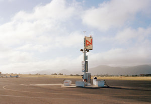 A photograph of an old airfield with a petrol filling station & old airplanes in the photo. Blue skies & clouds with palm trees in the background. USA