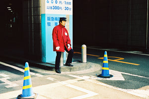 A parking attendant sitting outside a Japanese carpark entrance. He wears a red uniform with a black cap in Toykp Japan.