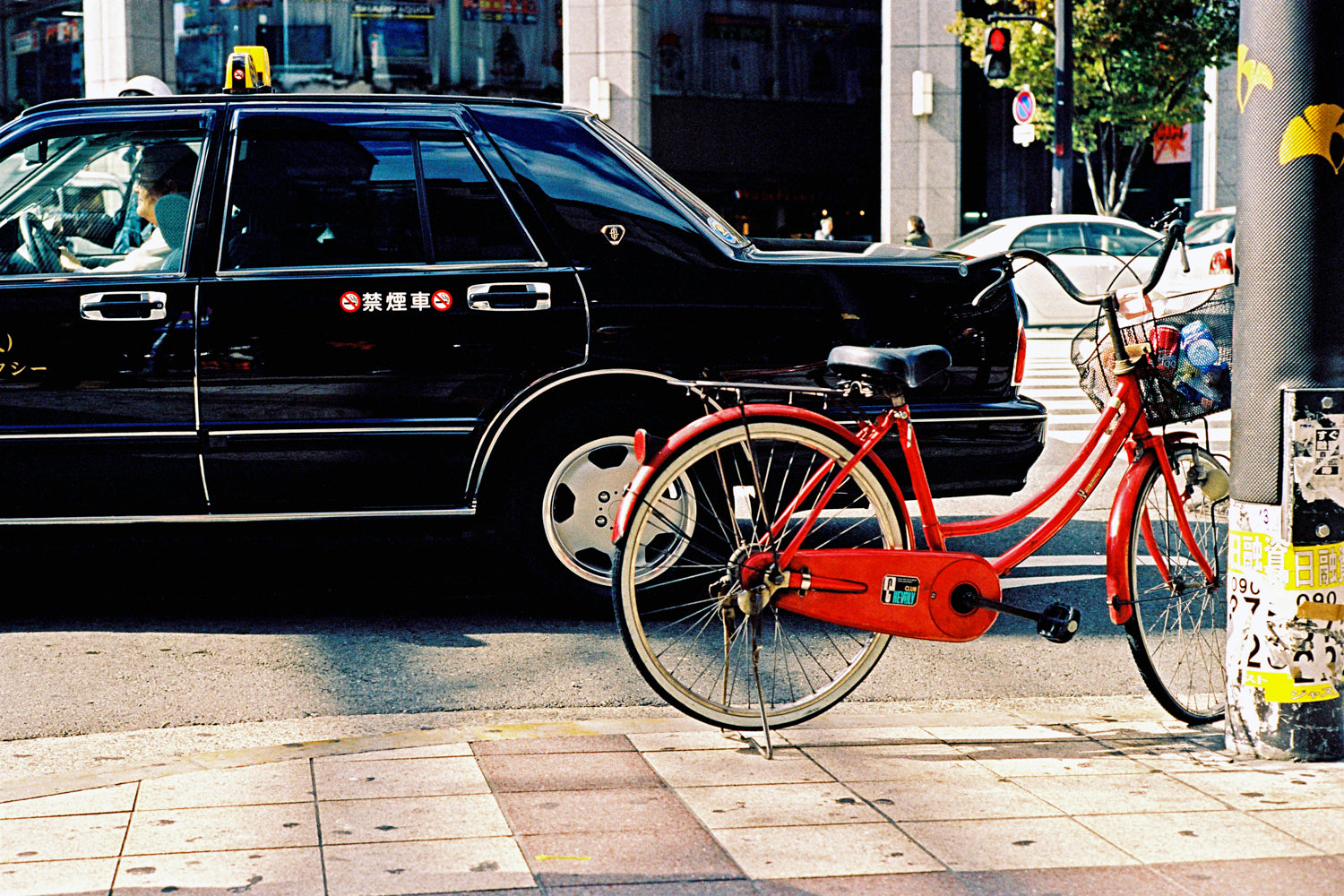 A photograph of a red bike leaning on a pole in a city street in Tokyo. There is a black taxi in the street.  Tokyo Japan. 