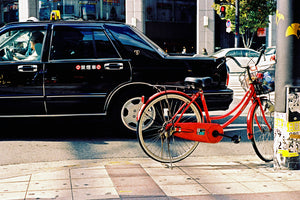 A photograph of a red bike leaning on a pole in a city street in Tokyo. There is a black taxi in the street.  Tokyo Japan. 