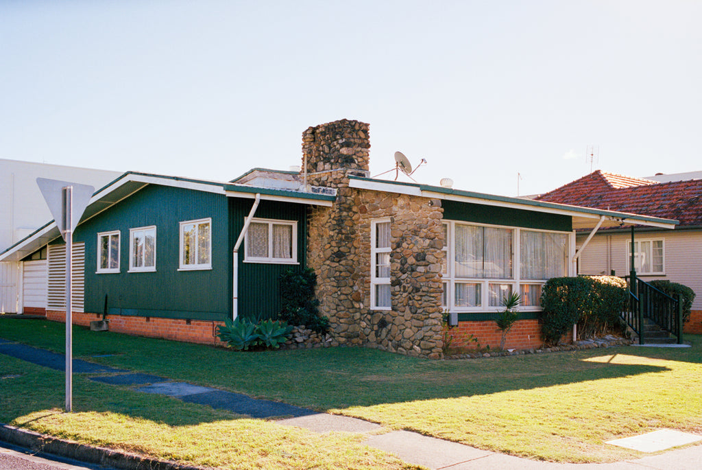 A photograph of a 1960’s timber house on the Goldcoast. The house is green with a large stone fireplace & chimney, blue skies.