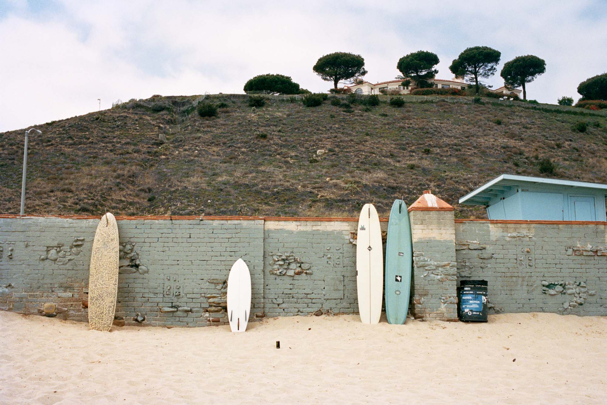 Surfboards lean on an old brick wall on Mailbu point surf break. There is a big hill with a house in the background