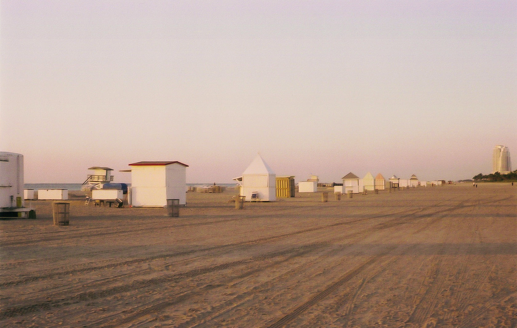 A photograph of an empty beach in Miami Florida. There are white beach boxes in the sand & the water is in the background.