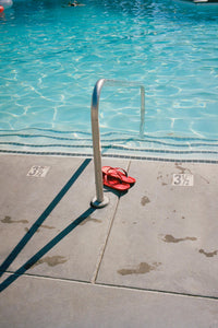 A photograph of a swimming pool with red flip flops on the edge of the pool. There is a marker in the concrete that says three & a half feet. The photograph is taken at the Ace Hotel on Palm Springs.